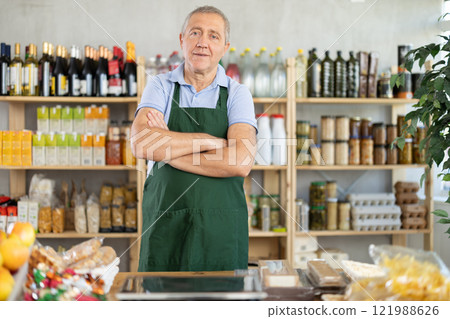 Elderly man in green apron stands near counter, works as salesman in grocery store. 121988626