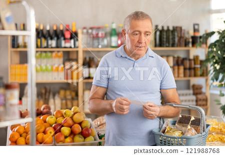 Elderly man with paper list in hands, walks around minimarket, puts groceries in market basket 121988768