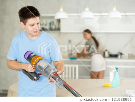 Young boy vacuum-cleaning in the kitchen while his girlfriend is dusting the kitchen 121988995