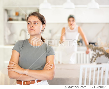 Young girl standing at the table while woman is quarreling with her standing behind Young girl standing at the table while woman is quarreling with her standing behind 121989083