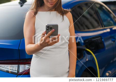 Woman uses smartphone while charging electric car in urban setting during daylight hours. Woman uses smartphone while charging electric car in urban setting during daylight hours. 121989777