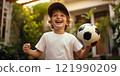 A cheerful young boy wearing a cap joyfully holds a soccer ball in his hands. The bright background features lush greenery, perfect for an outdoor sports theme. A simple and natural style embodies 121990209