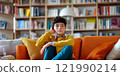 A thoughtful boy sits on a vibrant orange couch in a cozy living room. Shelves filled with books serve as a backdrop. This style captures the essence of relaxation and curiosity. AI 121990214
