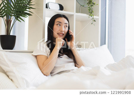 Relaxed Asian woman on bed having phone call. Young adult enjoying conversation in cozy home setting. Bright room enhances calm atmosphere. 121991908