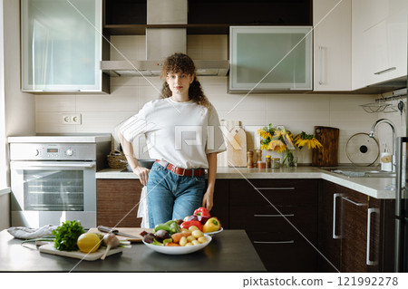 Woman stands in a kitchen surrounded by fresh fruits and vegetables on the table 121992278
