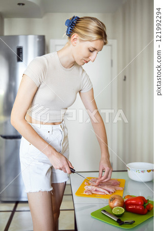 Young woman slices chicken on a bright yellow cutting board, surrounded by colorful vegetables Young woman slices chicken on a bright yellow cutting board, surrounded by colorful vegetables 121992294