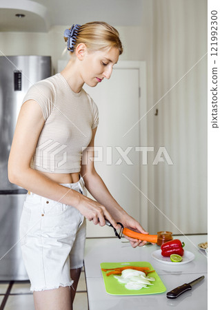 A young woman is peeling fresh carrot in a bright kitchen while cooking a healthy meal A young woman is peeling fresh carrot in a bright kitchen while cooking a healthy meal 121992300