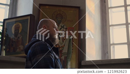 Man praying near golden altar inside orthodox church. Peaceful moment of faith and reflection, surrounded by icons and lit candles, symbolizing spirituality and devotion. Man crossing himself 121992623