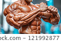 Close-up of a very muscular man's torso. Biceps, triceps, shoulders and abs are visible. The muscles are very developed and well-defined, the skin is shiny, probably from oil or sweat. 121992877
