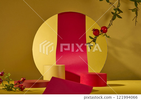 On a yellow background, a red paper roll is seen in front through a circle-shaped cutout. On a gold surface are two empty podiums for showcasing products. Some pomegranate branches as decorations. 121994066