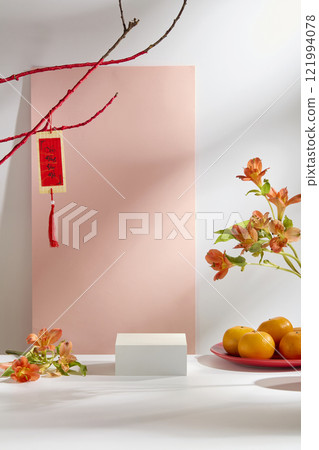 The New Year theme is set against a white backdrop featuring a blush pink panel. A cube as a product platform, decorated with red branches, calligraphy, alstroemeria, and a mandarin orange plate. 121994078