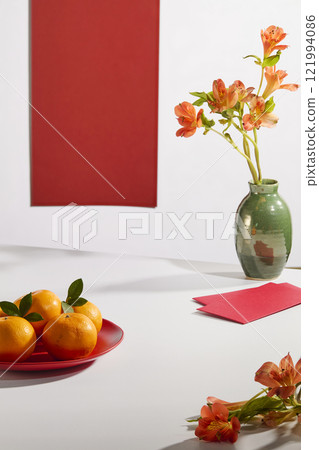 Front view of an empty space for displaying products, next to a vase of alstroemeria, mandarin oranges plates, red envelopes, and a red panel set in a white backdrop. Perfect for a New Year theme. 121994086