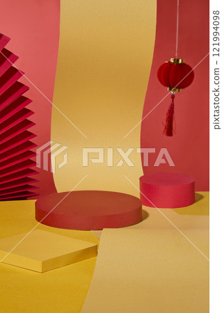 Against a red and gold paper backdrop. A curved gold paper as a focus, with cylindrical and square podiums to display products. A red pleated fan and a hanging red lantern add Lunar New Year detail. 121994098