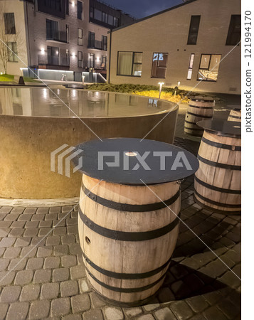 Wooden barrel tables with black tops sit beside a circular water feature at dusk. High quality photo 121994170