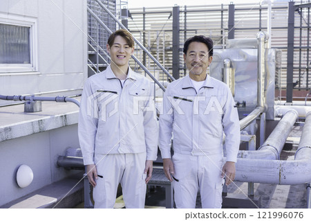 Two men in work clothes standing in front of the water supply and drainage equipment on the roof. Photo courtesy of Tokyo Electronics College, Denpa Gakuen Corporation. 121996076