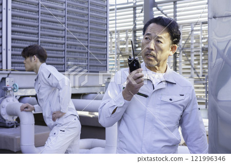 A man in work clothes talking on a walkie-talkie in front of the water supply and drainage equipment on the roof. Photo courtesy of Tokyo Electronics College, Denpa Gakuen Corporation. A man in work clothes talking on a walkie-talkie in front of the water supply and drainage equipment on the roof. Photo courtesy of Tokyo Electronics College, Denpa Gakuen Corporation. 121996346