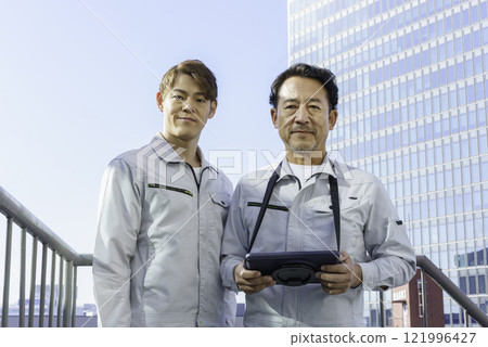Two men in work clothes standing with tablets on the roof of a building. Photo courtesy of Tokyo Electronics College, Denpa Gakuen Corporation. 121996427