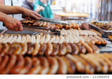 Close-up of assortment juicy bratwurst sausages and meat steaks cooking on grill at German street food market fest. Assorted grilled sausages on barbecue  at an outdoor streetfood marketstal 121996959