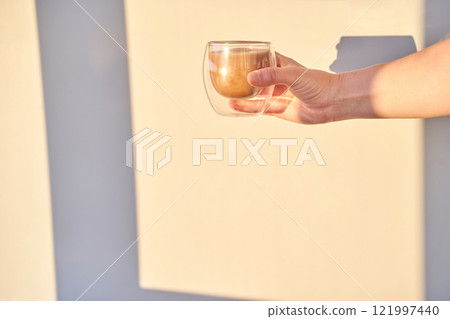 Female hand holding glass cup of coffee against sunlit beige wall in minimalist 121997440