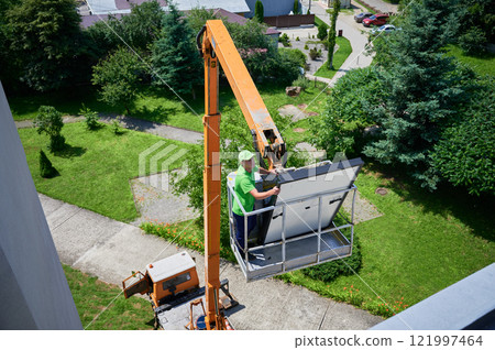 Workers lifting up photovoltaic solar panel on metal rooftop of house with assistance of crane lift. Men installers installing photovoltaic solar modules outdoors. Renewable energy generation concept. Workers lifting up photovoltaic solar panel on metal rooftop of house with assistance of crane lift. Men installers installing photovoltaic solar modules outdoors. Renewable energy generation concept. 121997464