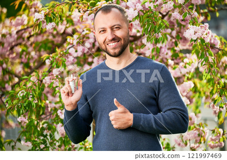 Man allergic using medical nasal spray, suffering from seasonal allergy at spring in blossoming garden. Handsome smiling man showing thumbs up near blooming tree outdoors. Spring allergy concept. Man allergic using medical nasal spray, suffering from seasonal allergy at spring in blossoming garden. Handsome smiling man showing thumbs up near blooming tree outdoors. Spring allergy concept. 121997469