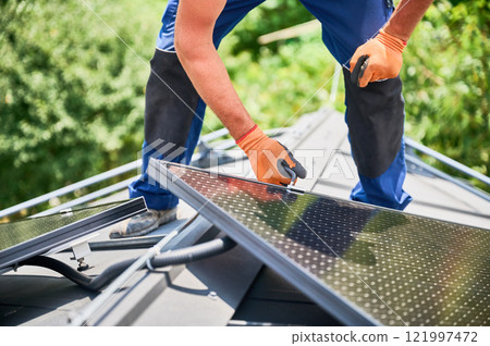 Worker building photovoltaic solar panel system on rooftop of house. Man engineer in gloves installing solar module with help of hex key outdoors. Alternative and renewable energy generation concept. Worker building photovoltaic solar panel system on rooftop of house. Man engineer in gloves installing solar module with help of hex key outdoors. Alternative and renewable energy generation concept. 121997472