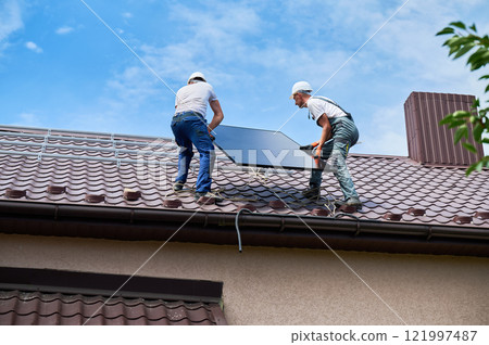 Workers building solar panel system on roof of house. Two men installers in helmets carrying photovoltaic solar module outdoors. Alternative, green and renewable energy generation concept. Workers building solar panel system on roof of house. Two men installers in helmets carrying photovoltaic solar module outdoors. Alternative, green and renewable energy generation concept. 121997487
