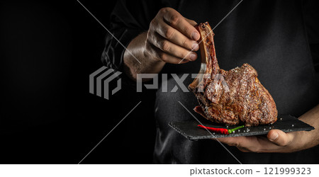 Chef hands holds Grilled Tomahawk Steak on bone on a dark background, Long banner format. copy space for text Chef hands holds Grilled Tomahawk Steak on bone on a dark background, Long banner format. copy space for text 121999323