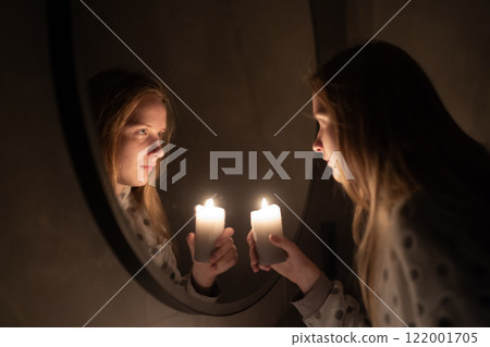 A young woman gazes into a mirror while holding a lit candle, the warm glow illuminating her face and its reflection. The serene moment evokes introspection and quiet elegance 122001705