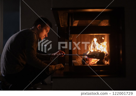 A young man tends to a fireplace, carefully adjusting a log with a fire poker. The warm glow of the flames illuminates his focused expression, creating a cozy and reflective atmosphere. 122001729