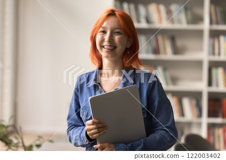 Successful stylish female teenager student posing in library holding laptop 122003402