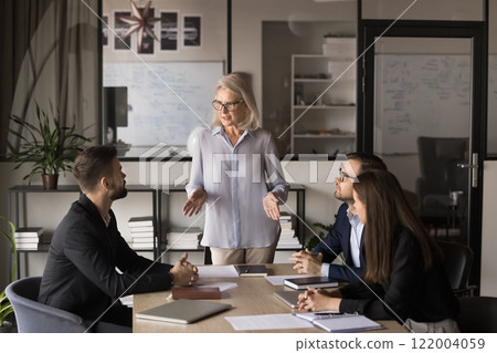 Businesswoman standing and addressing group of professionals seated at table 122004059