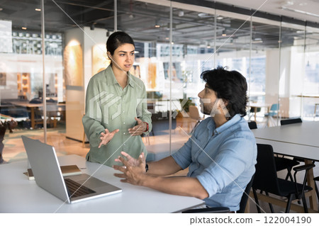 Two serious busy young Indian professionals talking at workplace Two serious busy young Indian professionals talking at workplace 122004190