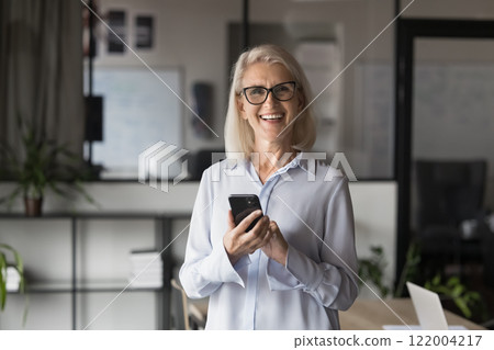 Positive middle-aged businesswoman posing at workplace with smartphone 122004217