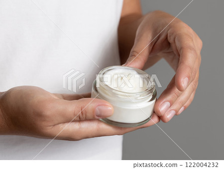 Female hand holding opened face cream jar against white t-shirt close up. Beauty concept Female hand holding opened face cream jar against white t-shirt close up. Beauty concept 122004232