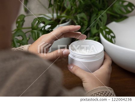 Woman in brown bathrobe touching cream with finger and holding a cosmetic jar in bathroom Woman in brown bathrobe touching cream with finger and holding a cosmetic jar in bathroom 122004236
