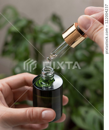Closeup of woman hands opening green dropper bottle aginst green plant, drop falling from pipette Closeup of woman hands opening green dropper bottle aginst green plant, drop falling from pipette 122004237