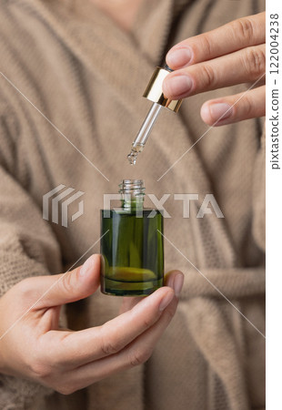 Closeup of woman in brown bathrobe opening green dropper bottle, cosmetic mockup Closeup of woman in brown bathrobe opening green dropper bottle, cosmetic mockup 122004238
