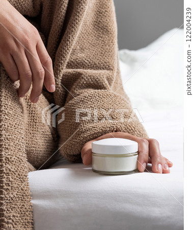 Woman in brown bathrobe puts white glass cream jar on white bedsheet closeup, cosmetic mockup 122004239