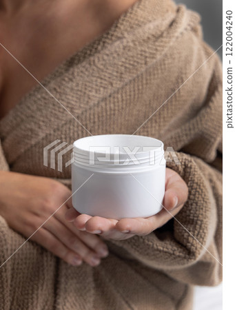 Closeup of woman in brown bathrobe holding opened blank cream jar in hands, cosmetic mockup 122004240