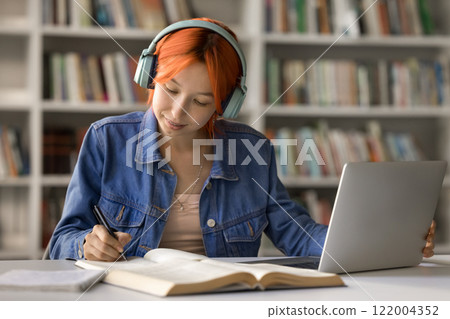Young female student in headset sitting by notebook taking notes Young female student in headset sitting by notebook taking notes 122004352