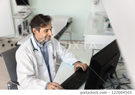 Positive Latin doctor man working at computer in hospital room 122004503