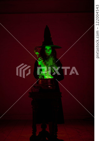 Mysterious witch in pointed hat, performing spell over glowing cauldron with hand sticking out, filled with green mist, surrounded by eerie red lighting. 122004543