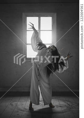 Exorcism. Monochrome creepy image of woman with black hair, in white gown with long claws stands barefoot in a creepy empty room Exorcism. Monochrome creepy image of woman with black hair, in white gown with long claws stands barefoot in a creepy empty room 122004561