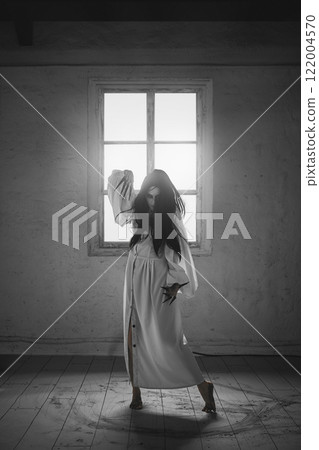 Powerful, mysterious woman, witch in white dress, with long black hair and claws stands in empty room with window and ash circles on floor. 122004570