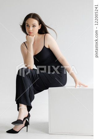 Fashion model posing sitting on white cube wearing black dress and high heels Fashion model posing sitting on white cube wearing black dress and high heels 122005241