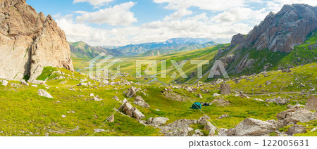 In the image, a green tent is set up at the foot of the cliffs under a blue sky with white clouds. Green grass and many stones typical of the nature of Kyrgyzstan are spread around In the image, a green tent is set up at the foot of the cliffs under a blue sky with white clouds. Green grass and many stones typical of the nature of Kyrgyzstan are spread around 122005631