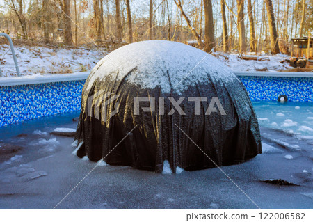 Snow covered pool cover rests in backyard surrounded by trees on cold winter day. Snow covered pool cover rests in backyard surrounded by trees on cold winter day. 122006582