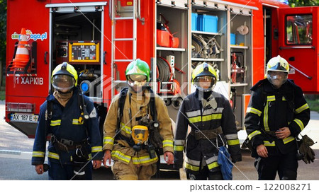 Portrait of male and female firefighters in helmets and protective uniforms standing near big red car. Young confident fireman and firewoman in full equipment posing against background of fire engine Portrait of male and female firefighters in helmets and protective uniforms standing near big red car. Young confident fireman and firewoman in full equipment posing against background of fire engine 122008271