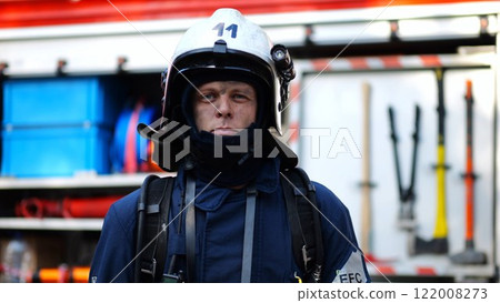 Portrait of confident male fireguard with dirty face standing near a fire engine. Young fireman in full equipment posing after fire against the background of big red truck. Concept of saving lives Portrait of confident male fireguard with dirty face standing near a fire engine. Young fireman in full equipment posing after fire against the background of big red truck. Concept of saving lives 122008273